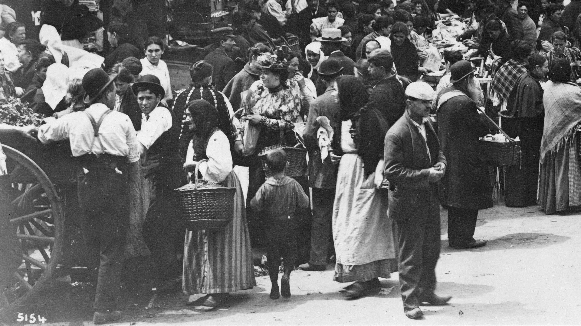 An enlarged section of the photo above. A woman in a fancy flowery blouse and ornate hat is purchasing goods at the center and all around her, people in more old fashioned clothing are doing the same.