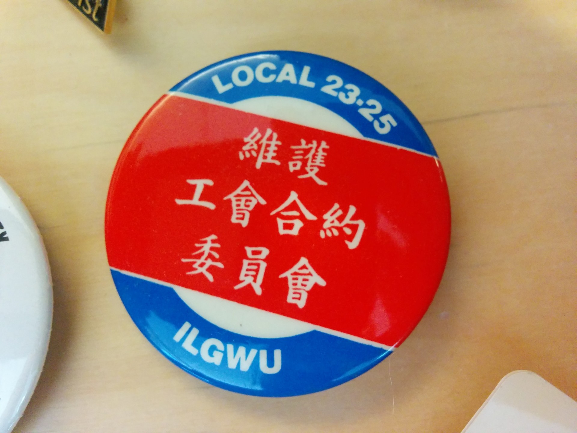 Close up on one circular union pin in red, white, and blue. The top of the pin reads, ‘Local 23-25’. The middle of the pin is three lines of Chinese. The bottom of the pin reads, ‘ILGWU’.