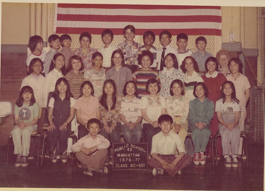 A diverse class of 31 students and one teacher pose for a photo. A sign in front reads, “Public School 42, Manhattan 1976-77, Class 6c-501.”