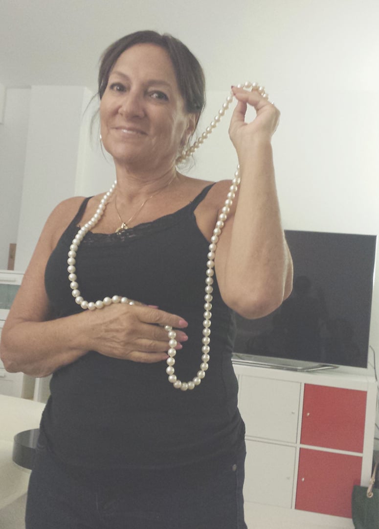 Bella as an adult wearing a black tank top and posing with a long string of pearls around her neck.