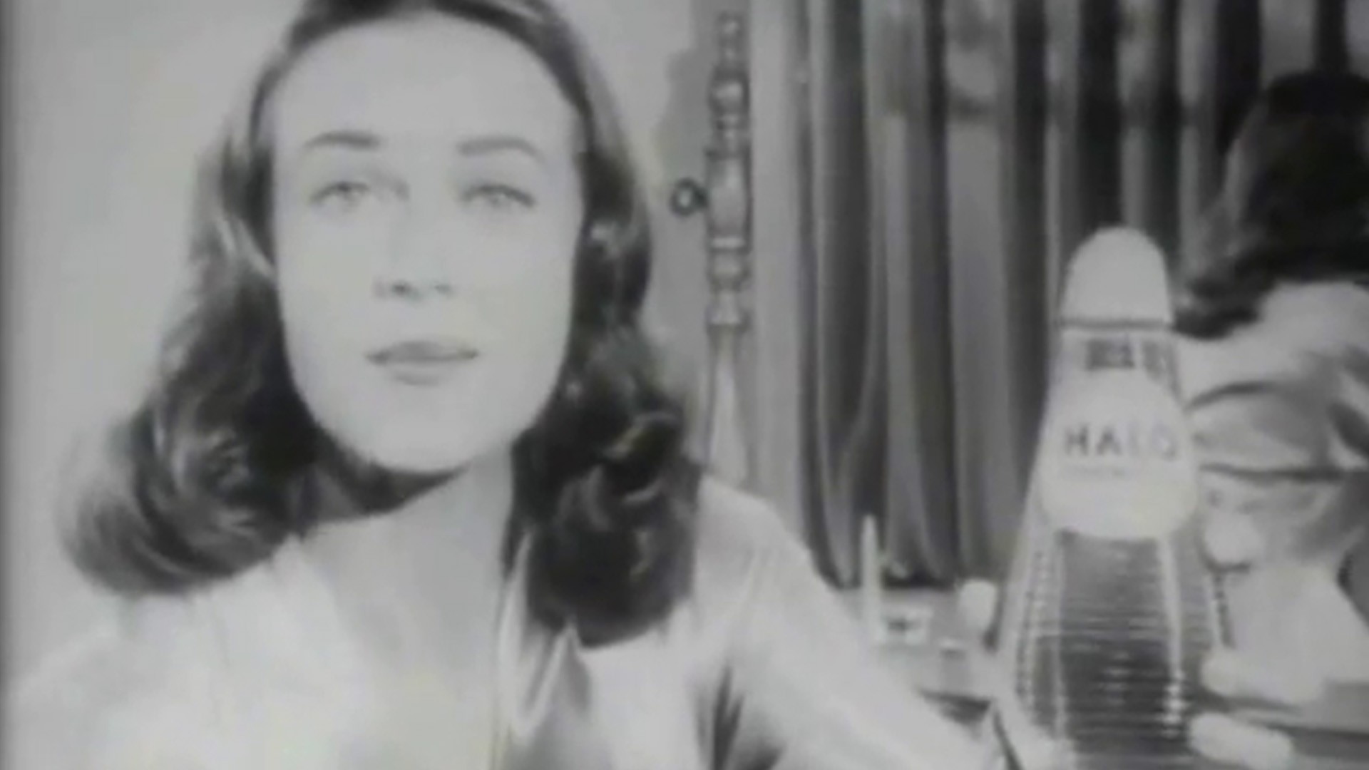 Black and white still image from a Halo Shampoo TV ad. Well-coiffed woman holding a bottle of Halo Shampoo.